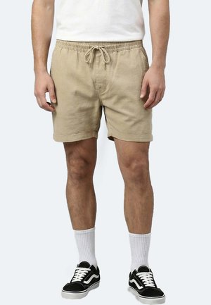 Mann trägt beige Shorts mit Kordelzug, weiße Socken, schwarz-weiße Turnschuhe und ein weißes T-Shirt und steht vor einem einfarbigen Hintergrund.
