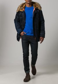 Puffer jacket with faux fur hood, navy color, zip and button closure, paired with a blue sweatshirt, dark jeans, and brown ankle boots.