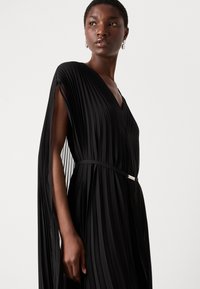 Black pleated dress with deep V-neck, flowing sleeves, and a slim belt with a silver accent. Fabric has a textured, layered look.