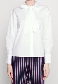 Blusa bianca di cotone con colletto a fiocco, maniche lunghe e chiusura a bottoni. Abbinata a pantaloni rigati blu navy con dettagli rosa.