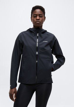 Γυναίκα που φοράει μαύρο φούτερ με κουκούλα και φερμουάρ και μαύρα κολάν, στέκεται μπροστά σε ένα απλό γκρι φόντο, τα χέρια χαλαρά στο πλευρό.