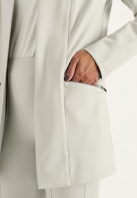 Ljusgrå blazer med en slät yta och en dragkedjeficka på sidan, i kombination med matchande figursydda byxor.