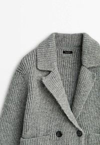 Blazer gris en tricot avec une texture côtelée, col cranté et deux boutons noirs. Comprend deux poches avant pour plus de détails.