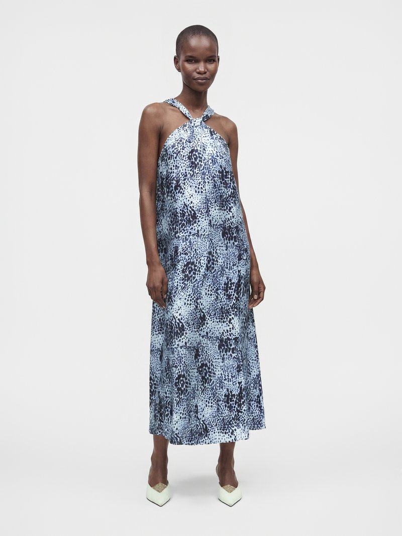 Femme portant une robe sans manches bleue à motifs avec un col halter et des chaussures blanches pointues, debout devant un fond clair uni.