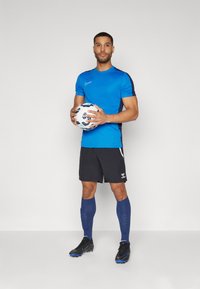 Kék, rövid ujjú sportpóló fekete oldalberakásokkal, fekete rövidnadrág, kék térd fölé érő zoknik és fekete stoplis cipők, egy mintás focilabdát tartva.