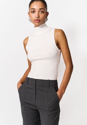 Femeie purtând un top alb fără mâneci cu guler înalt și pantaloni gri croiți, cu mâinile în buzunare și cercei mari rotunzi, pe fundal alb.