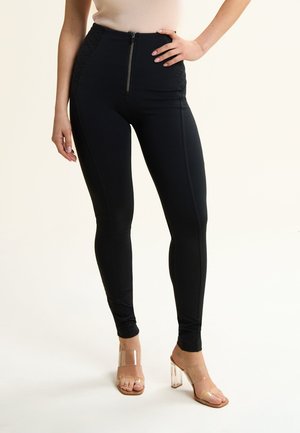 Leggings neri a vita alta realizzati in tessuto elastico, con una zip frontale e dettagli trapuntati sui lati. Sandali con tacco trasparente completano il look.