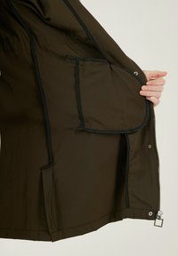 Jachetă de exterior maro, cu interior care prezintă buzunare cu margini negre. Fabricată dintr-un material neted, dispune de un fermoar și sistem de închidere cu capse.