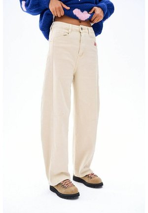 Osoba ubrana w luźne beżowe spodnie, brązowe buty trekkingowe z różowymi sznurówkami oraz niebieski sweter z różowymi wzorami serc, delikatnie podnosząca sweter.