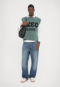 Groene gebreide vest met patroon en zwarte tekst, witte shirt eronder, lichtblauwe losse spijkerbroek en een zwarte schoudertas. Casual schoeisel.