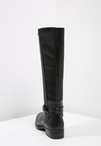 Botte haute en cuir noir avec un talon bas, détail de sangle à la cheville et texture lisse, présentée sur un fond blanc.