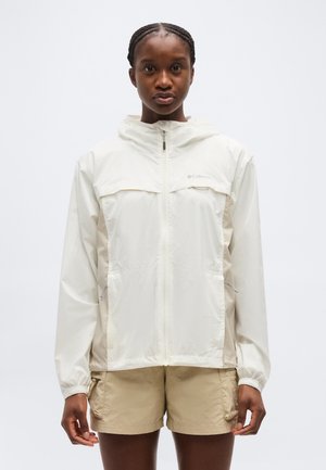 Punase palmikuga naine, kes kannab valget Columbia tuulekaitsejope ja beeži lühikesi pükse, seisab ühevärvilise tausta ees.