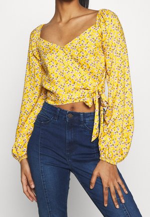 Femme portant un chemisier cache-cœur à manches longues jaune à motifs floraux, associé à un jean taille haute bleu foncé, main reposant sur la cuisse.