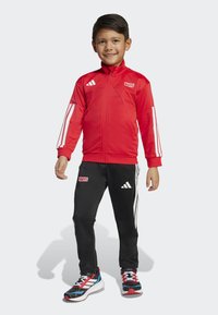 Rode sportjack met witte strepen, ritssluiting aan de voorkant en "MARVEL" logo. Zwarte trainingsbroek met witte strepen en "MARVEL" logo. Meerkleurige sneakers.