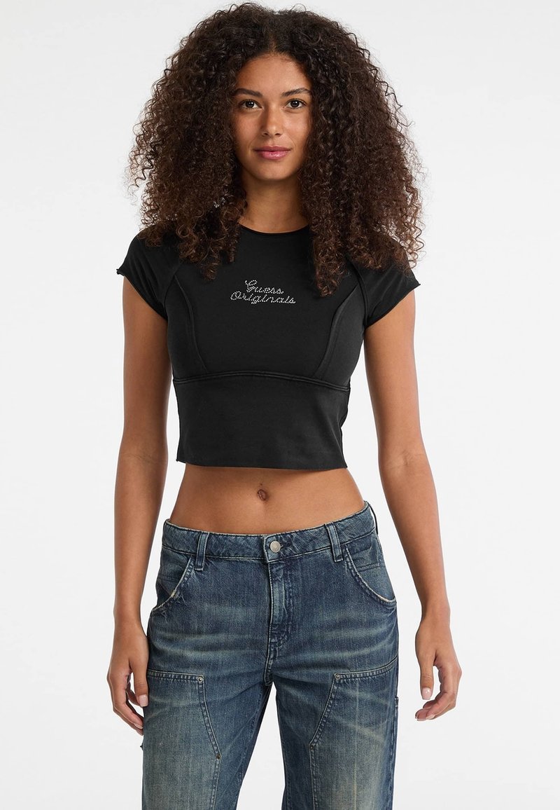Zwarte cropped t-shirt met korte mouwen, voorzien van een gestructureerd "Guess Originals" logo op de voorkant. Gecombineerd met vervaagde blauwe denim jeans.