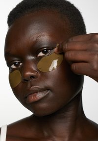 Le maschere in gel verde sono applicate sotto gli occhi di una persona con pelle scura. La pelle è liscia e lo sfondo è bianco semplice.