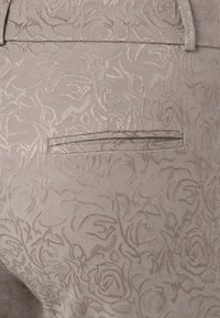 Tissu beige texturé avec un motif de rose en relief, comportant une poche arrière discrète. Design élégant avec des détails subtils.