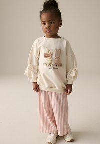 Cremefarbenes Sweatshirt mit zwei gestickten Hasen, die eine Schleife tragen, kombiniert mit rosa-weißen gestreiften weiten Hosen und weißen Sneakers.