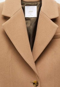 Manteau en laine couleur camel avec larges revers, un seul bouton visible, et une doublure intérieure marron avec une étiquette blanche de la marque au col.
