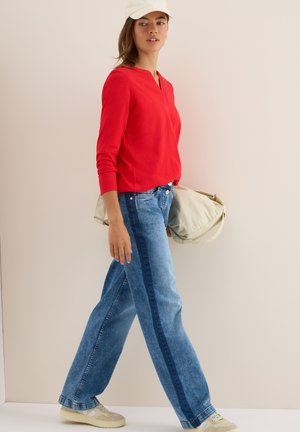 Femme marchant portant une chemise rouge à manches longues, un jean bleu bicolore, des baskets beiges, une casquette blanche et tenant un sac beige contre un fond uni.