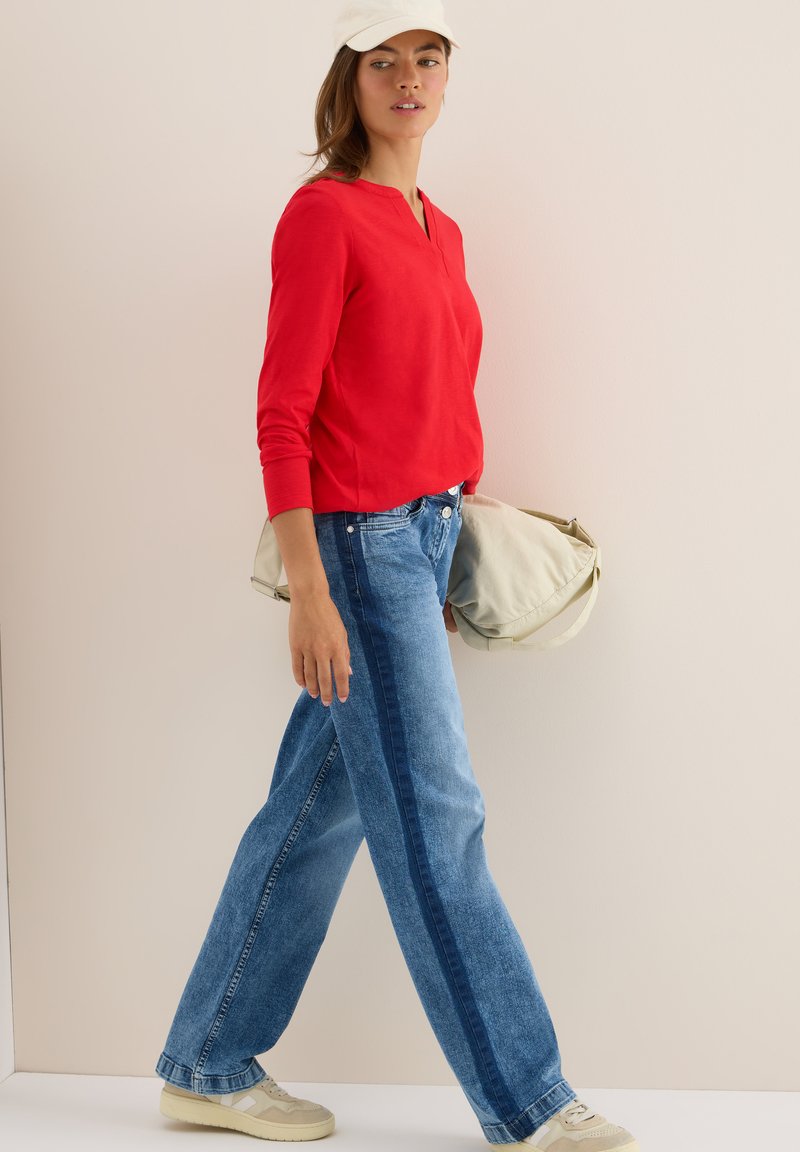 Kvinna går och bär en röd långärmad skjorta, tvåfärgade blåjeans, beige sneakers, vitkeps och håller en beige väska mot en enfärgad bakgrund.
