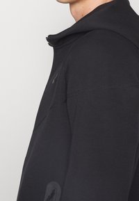 Svart zip-up hoodie med hög krage, slät textur och subtila sömdetaljer. Har en sidoficka med dragkedjestängning.