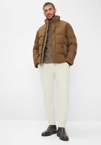 Giacca a vento marrone con colletto alto, chiusura con zip e tasche laterali. Indossata sopra un maglione a trama e abbinata a pantaloni chiari e stivali marroni.