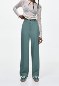 Pantalon large couleur sarcelle en tissu lisse, avec fermeture à boutons sur le devant et design plissé. Associé à des baskets vertes.