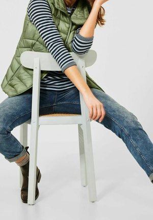 Persona seduta a ritroso su una sedia bianca, indossando un gilet imbottito verde, una maglia a righe con maniche lunghe, jeans blu e stivaletti neri alla caviglia.