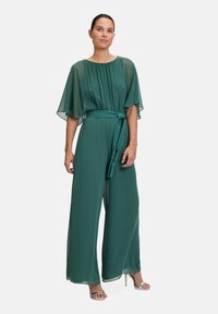 Tuta teal con maniche larghe, cintura cinchata e pantaloni lunghi flottanti. Realizzato in tessuto leggero e trasparente. Sandali con tacco argentati.
