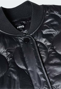 Czarna pikowana kurtka z dużymi okrągłymi szwami, prążkowanym kołnierzem i jednym widocznym czarnym zatrzaskiem z przodu.