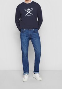 Felpa blu navy con logo grafico bianco, abbinata a jeans in denim blu e sneaker bianche. Outfit casual con una vestibilità comoda.