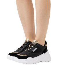 Baskets noires et dorées avec une semelle blanche épaisse, dotées de matériaux supérieurs texturés, d'accents en maille et d'un logo bien visible sur le côté.
