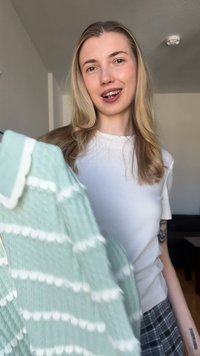 Mladá žena s dlhými blond vlasmi sa usmieva, drží zelený a biely pletený sveter v interiéri, oblečená v bielej košeli a kárované sukni.