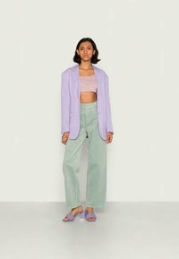 Mulher vestindo um blazer lavanda, uma blusa de flores lilás curta, calças largas verde menta e sandálias de salto lavanda, de pé contra uma parede lisa.