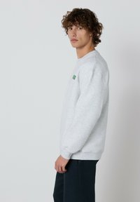 Mladý muž s kučeravými vlasmi, oblečený v svetlosivom mikinovom svete a tmavých nohaviciach, stojí v profilu proti jednoduchému svetlému pozadiu.