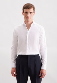 Camicetta bianca a maniche lunghe con colletto a punta, con chiusura a bottoni e una texture liscia, abbinata a pantaloni scuri. Design semplice e minimal.