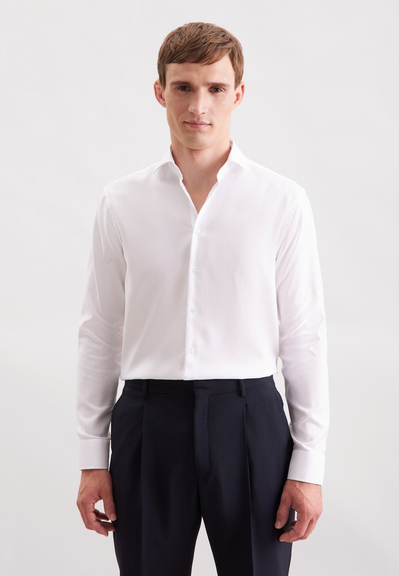 Camicetta bianca a maniche lunghe con colletto a punta, con chiusura a bottoni e una texture liscia, abbinata a pantaloni scuri. Design semplice e minimal.
