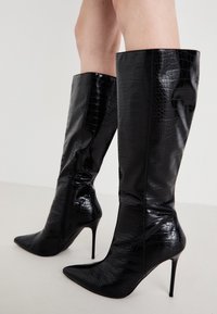 Svarta lackläderknähöga stövlar med krokodiltextur, spetsig tå, hög stilettklack och sidodetalj med dragkedja.