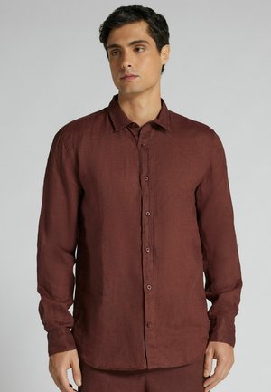 Ung mand iført en langærmet, knappet, mørkerødbrun skjorte med krave, stående mod en ensfarvet lys grå baggrund.