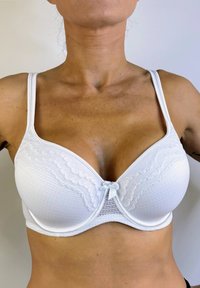 Sostenedor de encaje blanco con un diseño estructurado de aros, tirantes ajustables y un detalle de lazo en el centro. Textura de tela suave.