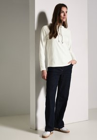 Sudadera blanca texturizada con cordón y bolsillo frontal, combinada con pantalones vaqueros azul oscuro y zapatos blancos sobre un fondo claro.