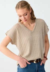 Femme portant un haut sans manches beige en maille côtelée avec col en V, jean bleu, ceinture marron avec boucle dorée, mains dans les poches, regard vers le bas.