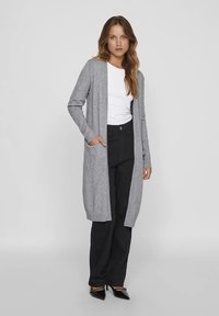 Mulher vestindo um longo cardigan cinza sobre uma blusa branca e calças pretas de cintura alta, de pé com uma mão no bolso.