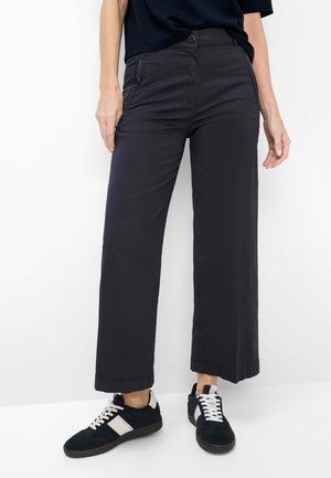 Kvinde iført sorte bukser med høj talje og vide ben samt sorte sneakers med hvide snørebånd og sideløbende striber, stående mod en hvid baggrund.