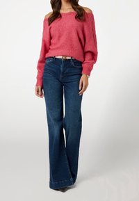 Pull rose à épaules dénudées avec tricot texturé, assorti à un jean bleu évasé. L'ensemble comprend une ceinture de couleur claire et des chaussures à talons.
