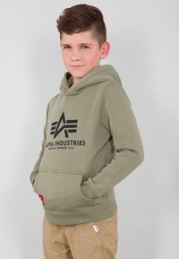 Olivově zelená mikina s kapucí a přední kapsou, s černým logem "Alpha Industries". Materiál je měkký, s šňůrkou pro stažení kapuce.