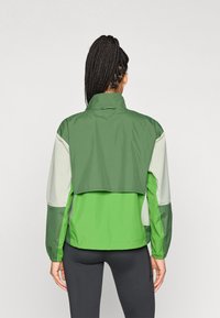 Chaqueta cortavientos verde y gris claro con cuello alto, puños elásticos y un diseño de espalda corta. Material ligero y impermeable.