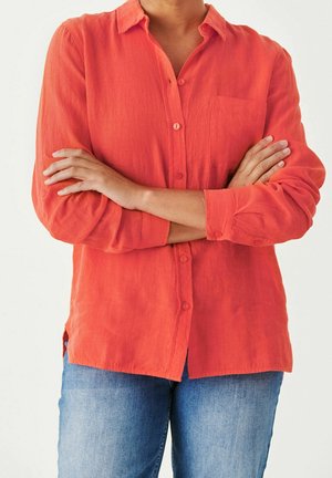 Person mit verschränkten Armen, trägt ein langärmeliges orangefarbenes Hemd mit Knöpfen und blaue Jeans, steht vor einem schlichten Hintergrund.