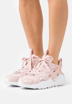 Person trägt blassrosa Nike-Sneaker mit weißen Sohlen und steht vor einem weißen Hintergrund.